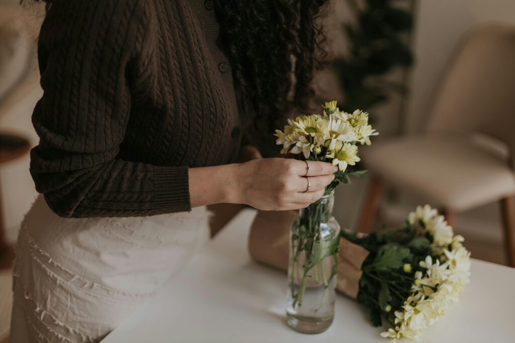 A woman arranges daisies in a glass vase indoors, creating a cozy atmosphere.